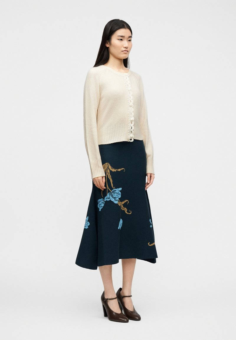 Vrouw die een crèmekleurig vest met knopen draagt, een donkerblauwe midi-rok met blauwe en bruine bloemrijke borduurwerk, en bruine Mary Jane schoenen met hoge hakken.