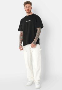 Schwarzes, übergroßes T-Shirt mit weißem Aufdruck "Sixty June" auf der Vorderseite, kombiniert mit lockeren weißen Hosen und weißen Sneakers. Tattoos sind auf den Unterarmen sichtbar.