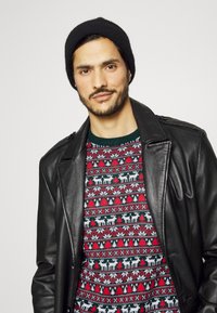 Črna usnjena jakna preko zeleni, rdeči in beli puloverja s potiskom jelena in dreves. Model nosi črno pleteno kapo.