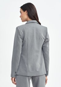 Szary marynarka o dopasowanym kroju z klapami, teksturowanym materiałem i mankietami na trzy guziki. Projekty ma wyraźnie zdefiniowaną sylwetkę.