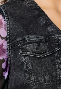 Czarna kamizelka dżinsowa z przednią kieszenią, teksturowaną tkaniną oraz okrągłym czarnym guzikiem z tłoczonymi detalami. Uzupełniona kwiatowym nadrukiem pod spodem.