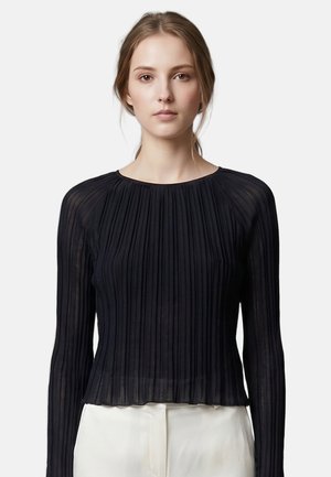 Jeune femme aux cheveux clairs attachés, portant un haut noir plissé à manches longues et un pantalon clair sur un fond uni.