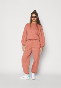 Sudadera de color naranja quemado y joggers a juego, hechos de material suave, con cintura con cordón y puños elásticos. Zapatillas blancas con detalles en azul.