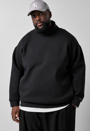 Mann trägt eine graue New York Yankees Baseballkappe, schwarzen Sweatshirt über weißem Hemd und schwarze Hose vor schlichtem hellem Hintergrund.