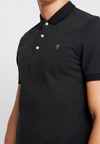 Man med en svart kortärmad polo skjorta med tre vita knappar och ett litet gult broderat "F" på bröstet.