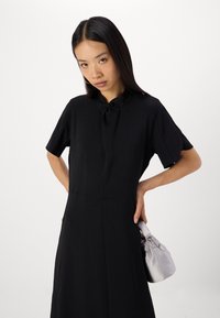 Schwarzes Kleid mit kurzen Ärmeln und einem hohen Ausschnitt, der mit einem Knotendetail versehen ist. Das Model hält eine hellgraue Satin-Handtasche mit einem verdrehten Griff.