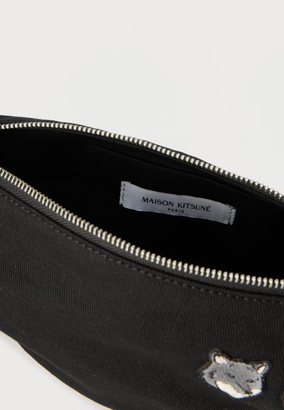 Trousse noire zippée avec un écusson de tête de loup et une étiquette intérieure indiquant "Maison Kitsuné Paris".