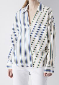 Femme portant une chemise blanche ample avec des rayures verticales bleues, vertes et jaunes, ainsi que un pantalon blanc, sur un fond uni.