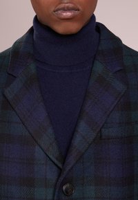 Blazer a quadri in misto lana verde scuro e blu navy, con rever a incavo, un bottone nero singolo e una tasca. Sotto, è visibile un maglione a collo alto.