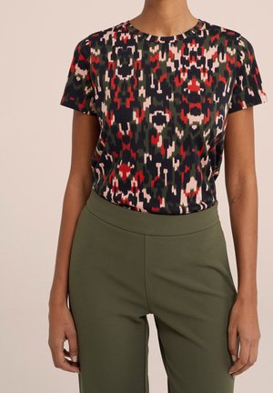 Osoba nosząca bluzkę z krótkim rękawem i okrągłym dekoltem, z abstrakcyjnym wzorem w kolorach czerwonym, czarnym, różowym i zielonym, wsuniętą w wysokie spodnie w oliwkowym kolorze.