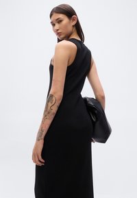 Μαύρο φόρεμα χωρίς μανίκια με εφαρμοστή σιλουέτα. Το μοντέλο κρατά μια μαλακή μαύρη τσάντα. Περιλαμβάνει διακριτική λάμψη και ομαλή υφή.