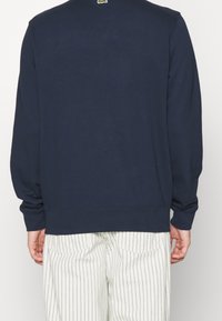 Sudadera azul marino con mangas largas, puños y dobladillo acanalados. Presenta un pequeño logotipo en la parte posterior del cuello. Combinada con pantalones blancos a rayas.