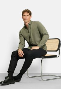 Mann mit lockigem Haar, der auf einem modernen Stuhl aus Rattan und Chrom sitzt. Er trägt ein olivgrünes Hemd mit Knöpfen, schwarze Hosen und schwarze Schuhe.