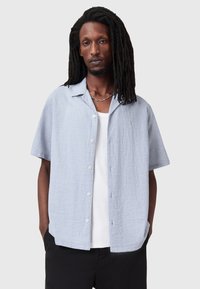 Camicia azzurra a maniche corte con tessuto texturizzato, chiusura con bottoni e colletto a punte, indossata sopra un top bianco.