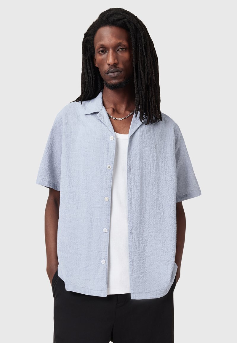 Camicia azzurra a maniche corte con tessuto texturizzato, chiusura con bottoni e colletto a punte, indossata sopra un top bianco.