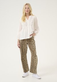Camicia bianca con bottoni e ricami, abbinata a pantaloni larghi della stampa leopardata marrone e sneaker bianche su uno sfondo neutro.
