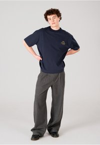 Camisa de hombre de gran tamaño en color navy con mangas cortas y cuello redondo, con un sutil logo bordado en dorado. Combinada con pantalones anchos grises.