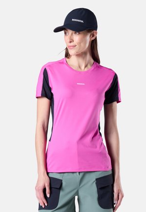 Femme portant un haut de sport à manches courtes rose vif et noir, un pantalon cargo vert, et une casquette noire Rossignol, regardant légèrement sur le côté.