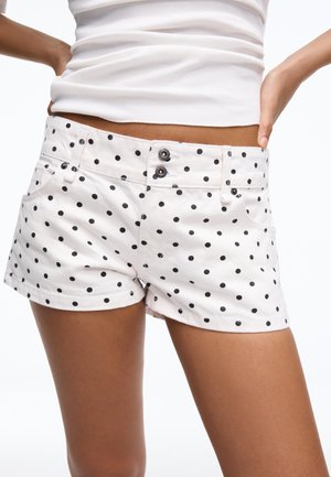 Hvide shorts med sorte polkaprikker båret af en person med hænderne på hoften, kombineret med en cropped hvid top.