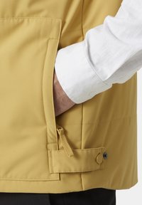 Chaleco amarillo de tejido suave, con un bolsillo lateral con cremallera y un cierre con botón para un ajuste ajustable. Manga de camisa blanca visible.