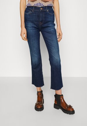 Tamsiai mėlynos džinsinės kelnės su tiesiu kirpimu ir neapdirbtu hemu, derinamos su rudo spalvos auliniais batais, akcentuotais grandinėmis ir masyviu padu.