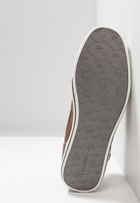 Semelle d'une chaussure marron avec une semelle extérieure en caoutchouc gris à motif géométrique et une semelle intermédiaire blanche, appuyée contre un mur blanc.