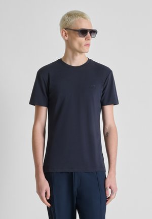 Uomo biondo che indossa occhiali da sole neri, una T-shirt nera a maniche corte semplice e pantaloni blu scuro, in piedi davanti a uno sfondo grigio uniforme.