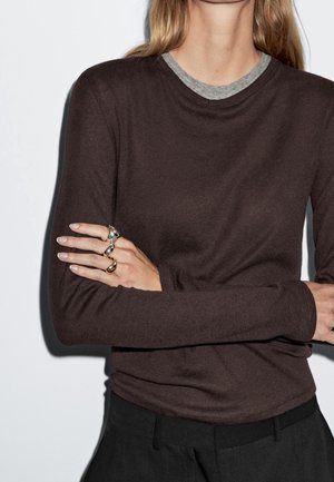 Donna con capelli biondi che indossa una camicia a maniche lunghe marrone scuro sopra una maglia grigia a girocollo, pantaloni neri e anelli d'argento e oro sulla mano sinistra.