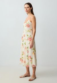 Robe florale crème, ornée de fleurs roses et de feuilles vertes. Silhouette évasée avec des bretelles fines et une jupe fluide, longue jusqu'au sol.