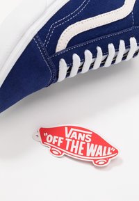 Baskets Vans en suède bleu avec des accents blancs, lacets et semelle en caoutchouc, accompagnées d'une étiquette rouge "VANS OFF THE WALL" sur un fond blanc.