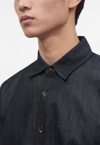 Jeune homme portant une chemise en denim foncé à boutons avec col, montré du cou jusqu'au milieu de la poitrine sur un fond clair uni.