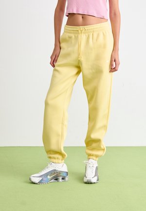 Osoba w żółtych spodniach dresowych Nike i białych butach Nike, stojąca na zielonej podłodze na tle jednolitego jasnego tła.