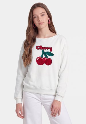 Ung kvinde med langt hår iført en hvid sweatshirt med røde paliet-kirsebær og ordet "Cherry" i rødt over dem.