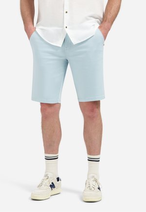 Man draagt lichtblauwe korte broek, wit shirt met handen in de zakken, witte sokken met zwarte strepen en witte sneakers met marineblauwe accenten.