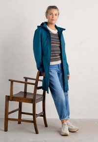 Teal vattentät jacka med huva, över en flerfärgad mönstrad tröja. Kombinerad med ljusblå jeans och vita sneakers, stående bredvid en trästol.