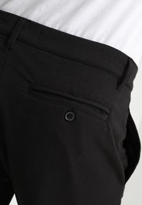 Pantalones negros con una textura suave, que cuentan con un bolsillo trasero con cierre de botón. La cintura es parcialmente visible, debajo de una camisa gris.