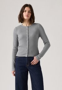 Cardigan grigio a coste con maniche lunghe, scollo rotondo e chiusura frontale con bottoni. Indossato sopra jeans scuri a vita alta in denim.