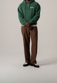 Hombre vestido con sudadera verde con el texto "BLKVIS", pantalones marrones, mocasines en blanco y negro, collar de oro y anillos, de pie con los brazos cruzados.