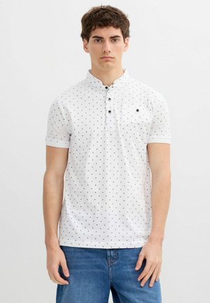 Joven con una camisa blanca de manga corta con pequeños puntos negros y vaqueros azules, de pie frente a un fondo liso.