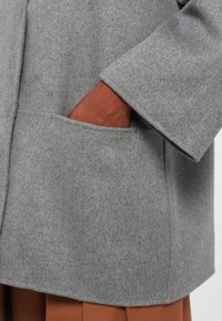 Mână în buzunarul unei lanterne din lână gri, arătând textura paltonului și detaliile mânecii peste pantaloni maronii.