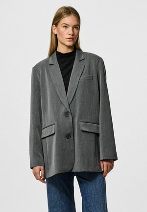 Blazer gris avec une coupe ample, doté d'une fermeture à deux boutons, de deux poches avant et d'un tissu texturé subtil. Associé à un haut sombre et à un jean large.