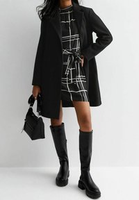 Cappotto nero su un vestito a scacchi bianco e nero con cintura in vita, abbinato a stivali neri al ginocchio. Piccola borsa nera con manici.