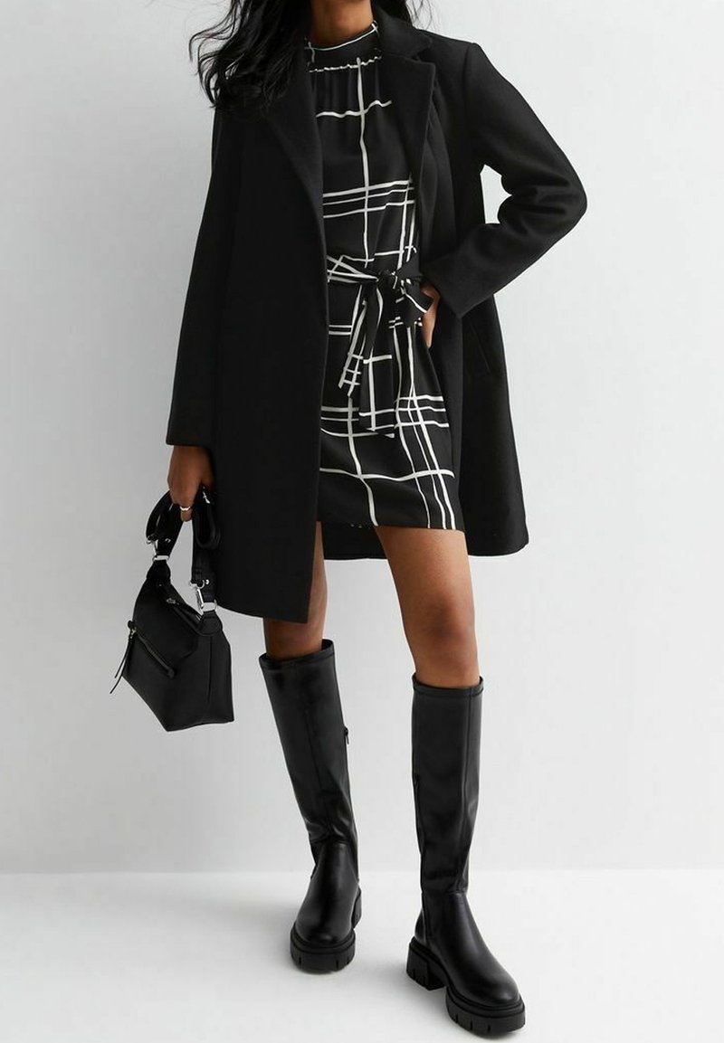 Cappotto nero su un vestito a scacchi bianco e nero con cintura in vita, abbinato a stivali neri al ginocchio. Piccola borsa nera con manici.