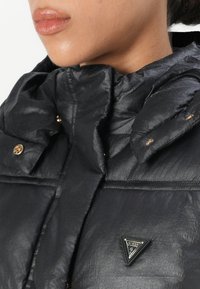Zwarte gewatteerde jas met een hoge kraag, voorzien van gouden hardware en een driehoekige logopatch. Glad, glanzende textuur met zichtbare naden.