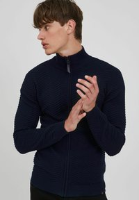 Mornarica zip-up dukserica s teksturiranim cik-cak uzorkom, visokim ovratnikom i rebrastim manžetama. Prilagođen dizajn koji naglašava oblik rukava i torza.