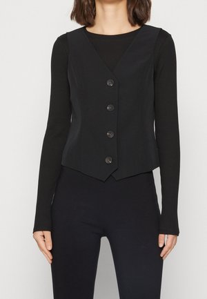 Vrouw die een zwart vest met knopen draagt over een zwart geribd shirt met lange mouwen en een zwarte broek, staand tegen een effen achtergrond.