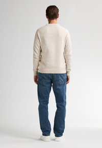 Beige gebreide trui met een ronde hals en ribgebreide zoom, gecombineerd met blauwe denim jeans en witte sneakers. Zichtbare stiksels op de jeans.