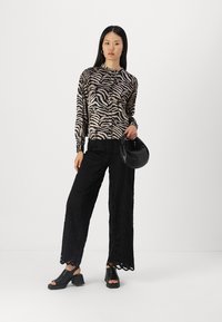 Juoda ir smėlio spalvos zebra raštuotas ilgomis rankovėmis palaidinė, derinama su permatomomis juodomis plačiomis kelnėmis su gėlių raštais. Modelis laiko juodą rankinę.