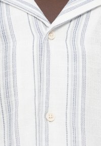 Άσπρο πουκάμισο με κάθετες ναυτικές ρίγες, μαλακό ύφασμα με υφή και άνετο γιακά. Διαθέτει τέσσερα κουμπιά μπροστά.