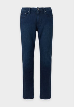 Jeune homme debout portant un t-shirt blanc à manches courtes, un jean bleu foncé et des baskets noires à semelles blanches devant un fond uni.